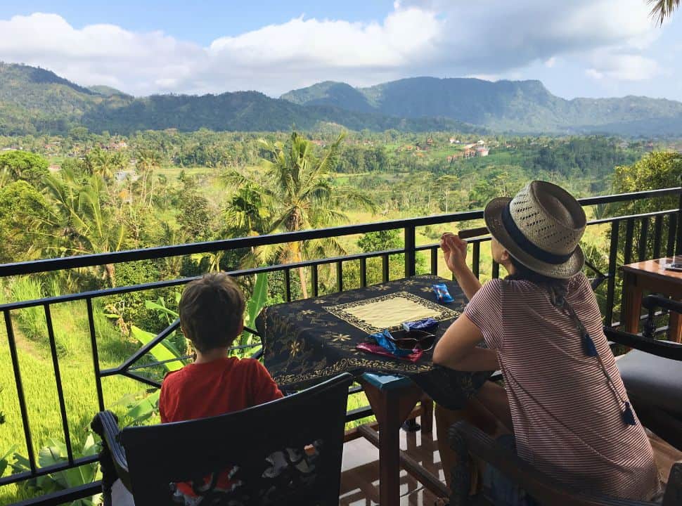 mother and son eating ice cream on a balcony with view of the valley with rice fields, palmtrees and mountains beyond in Sidemen Bali