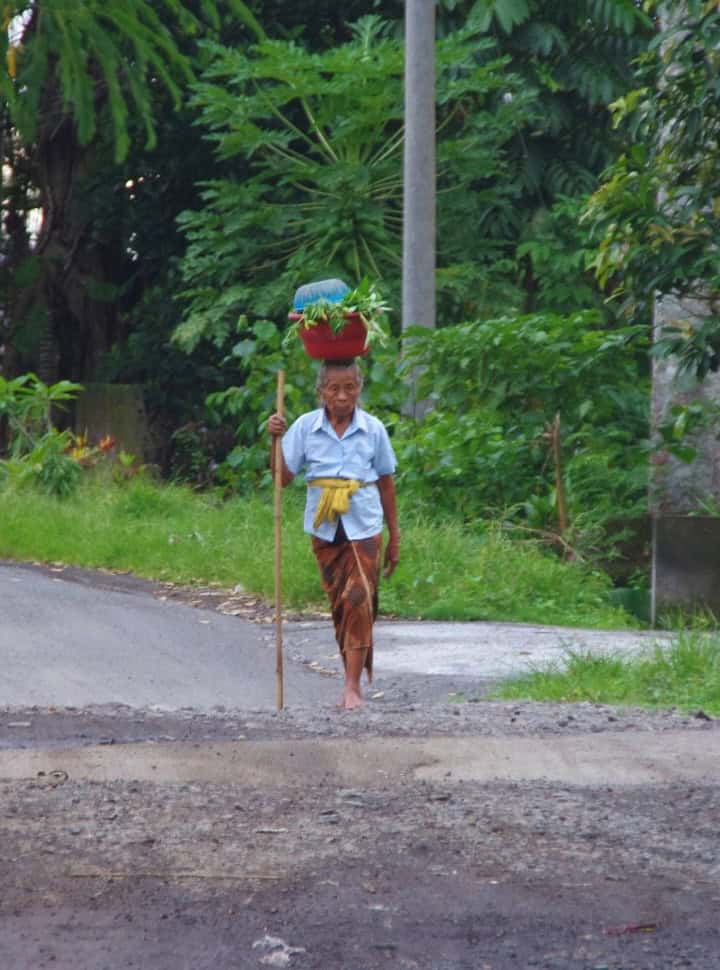 woman balancing vegetables on her head while holding a stick to aid her with walking barefoot along the small road in Sidemen