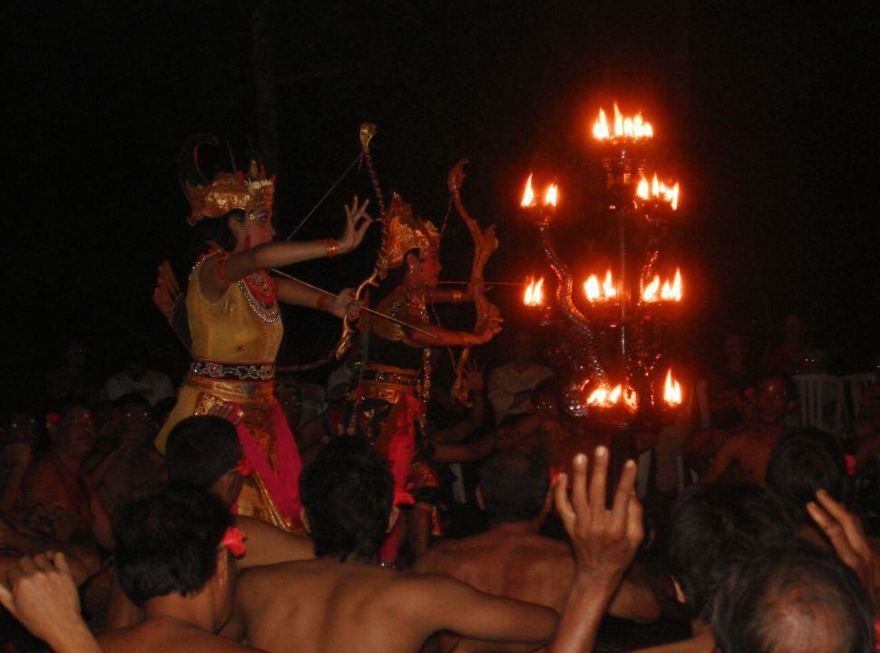 two Balinese dancers around a tall torch while being surrounded by kecak singers, a traditional Balinese dance
