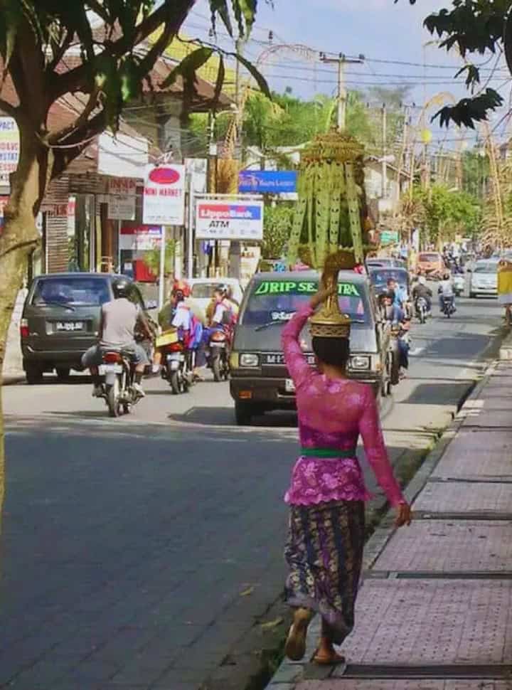 woman dressed in beautiful traditional clothing balancing a high offering on her head while walking down the busy road with traffic in Ubud Bali
