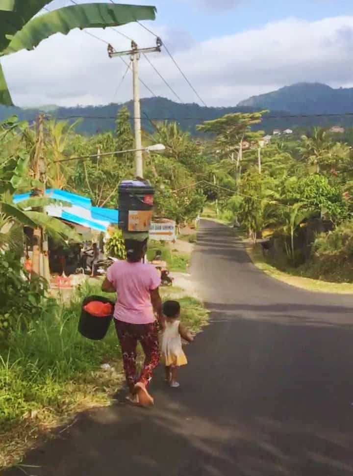 mother carrying laundry in a bucket and balancing on her head while a child walks in front of her at one of the many little streets in Sidemen