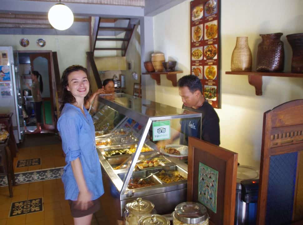 woman posing in a restaurant where various dishes are served buffet style, a man is placing various dishes on a place with rice