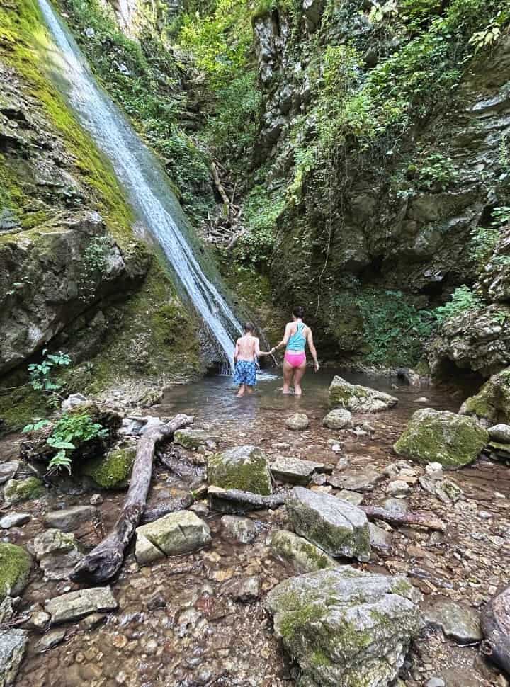mother and son wading into a waterfall pool which is surrounded by rocks and plants in slovenia