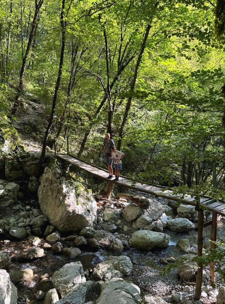 father and son standing on a wooden bridge above a shallow river with lots of rocks in a green forest, on their way to the waterfall in slovenia