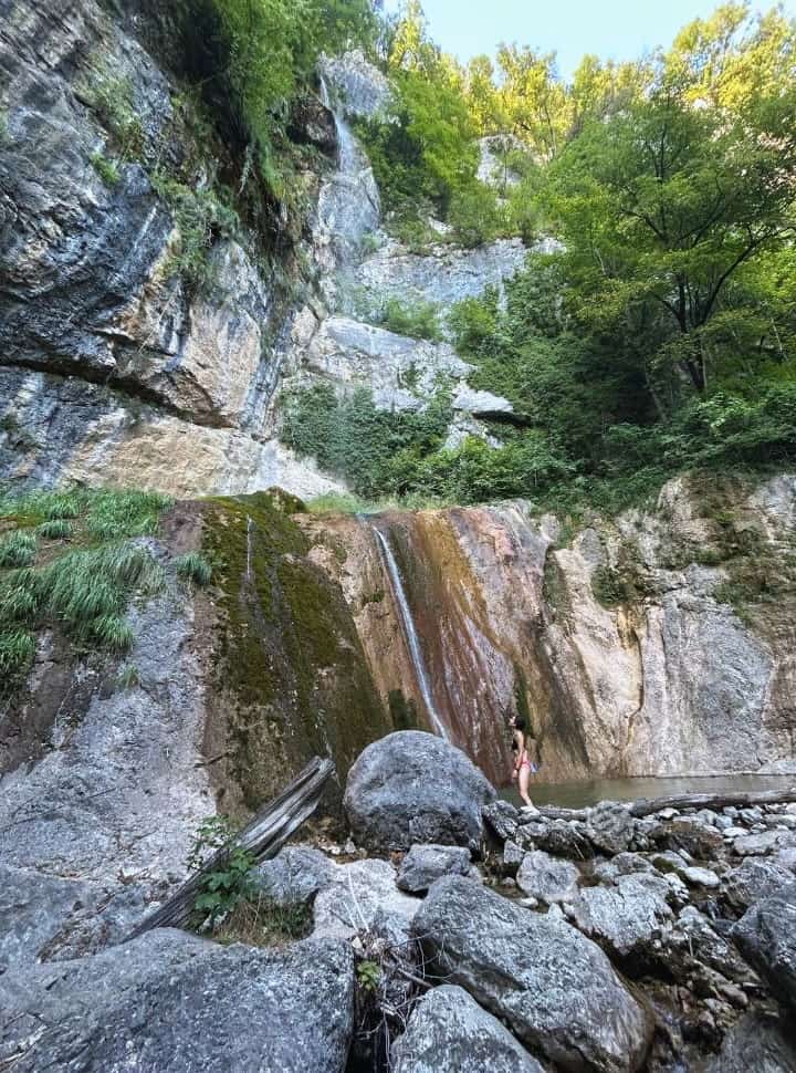 woman standing on the bottom of the waterfall surrounded by rocks and vegetataion