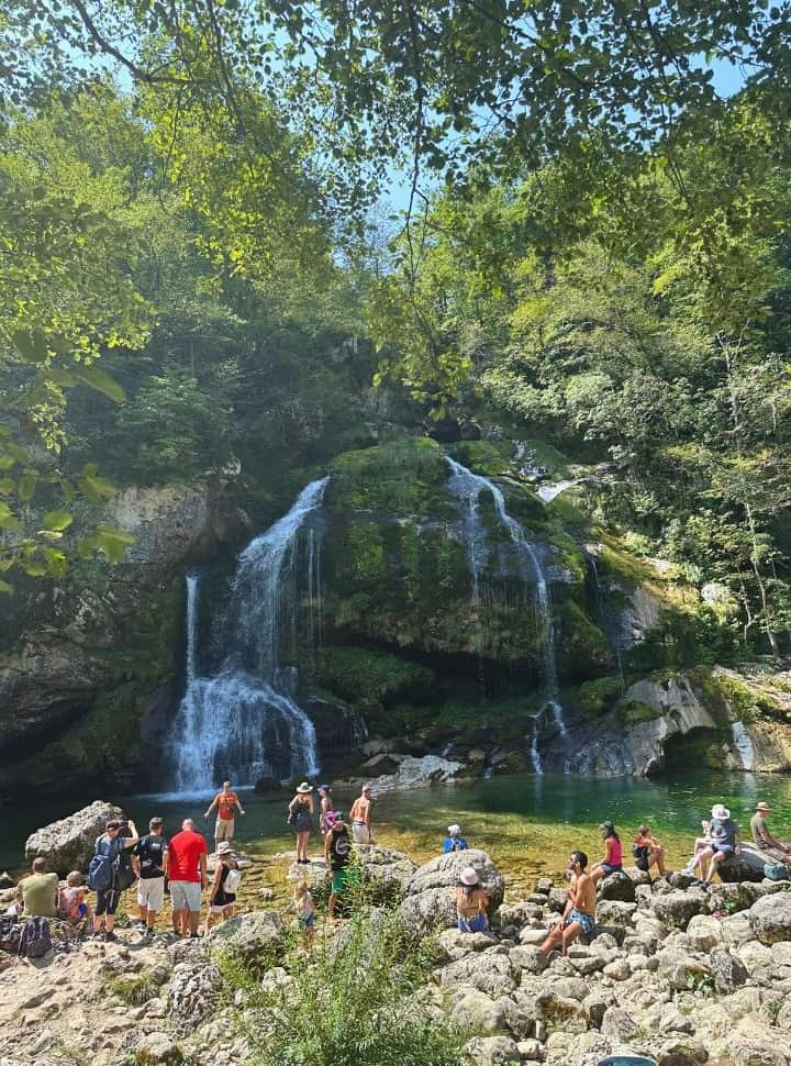 large group of people sitting on the rocks on the edge of a large waterfall pool admiring the waterfall and the natural setting at Virje Waterfall Slovenia