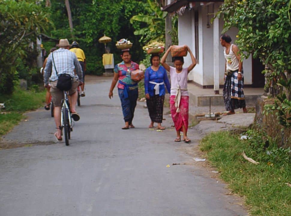 man on a mountain bike passing a village where three ladies are carrying baskets on their heads while walking along the small road