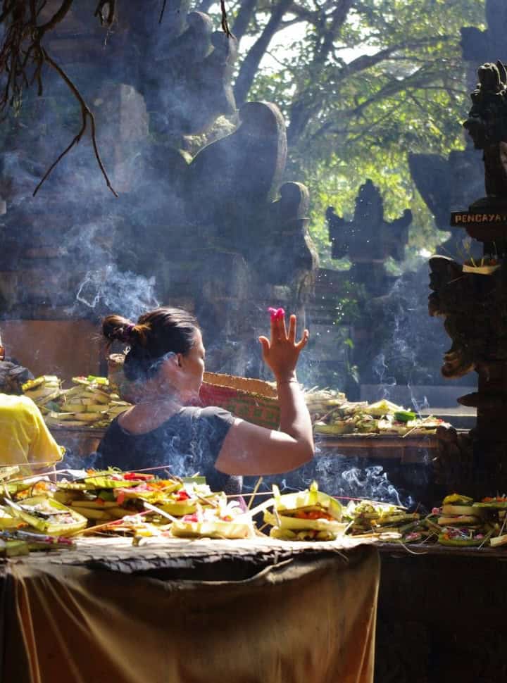 Balinese woman standing within a Balinese temple shrines, placing an offering with hundreds of others