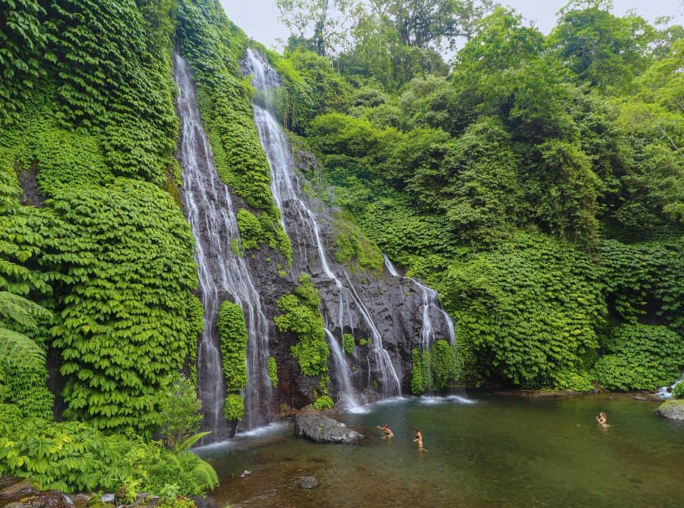 impressive tall waterfalls plunging into a large pool where people are swimming nearby Munduk Bali