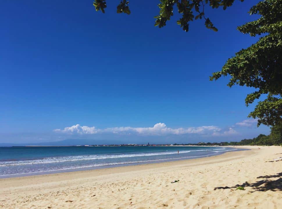 long stretched beach if soft sand, calm waves and blue water. A string of trees grow along the water at jimbaran beach Bali