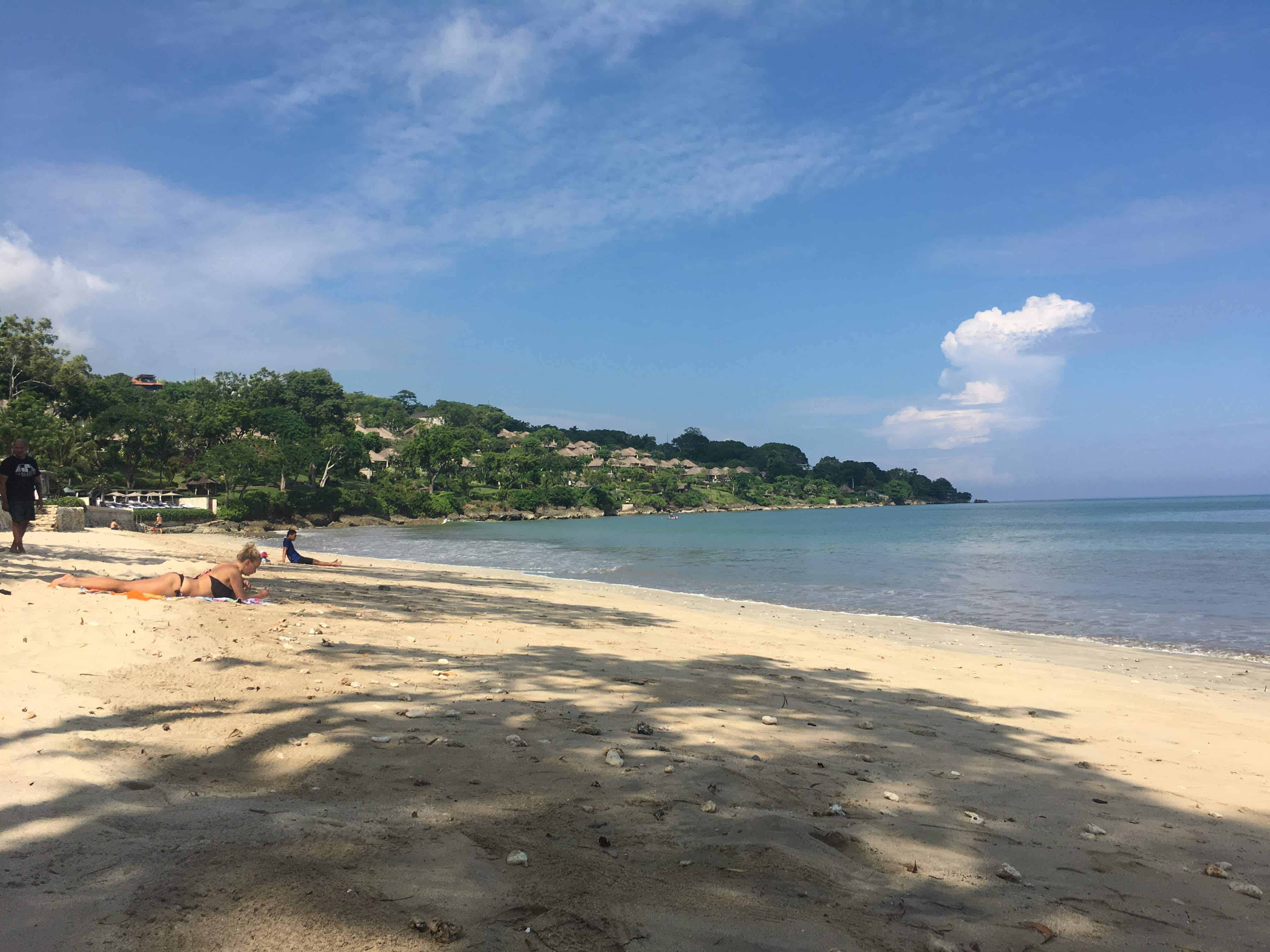 long stretched beach with soft sand, people are enjoying the beach while in the back there is accommodation on the hills