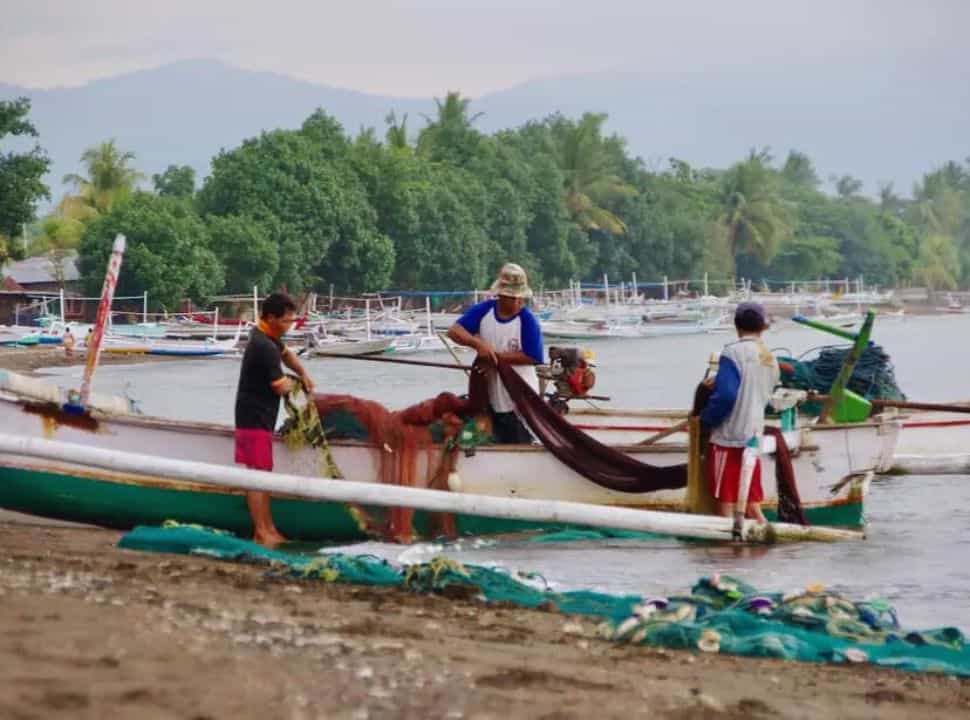 three fishermen at their traditional boat arranging their fishing nets, in the back there is a row of similar boats docked along the sea shore on the beach of Lovina