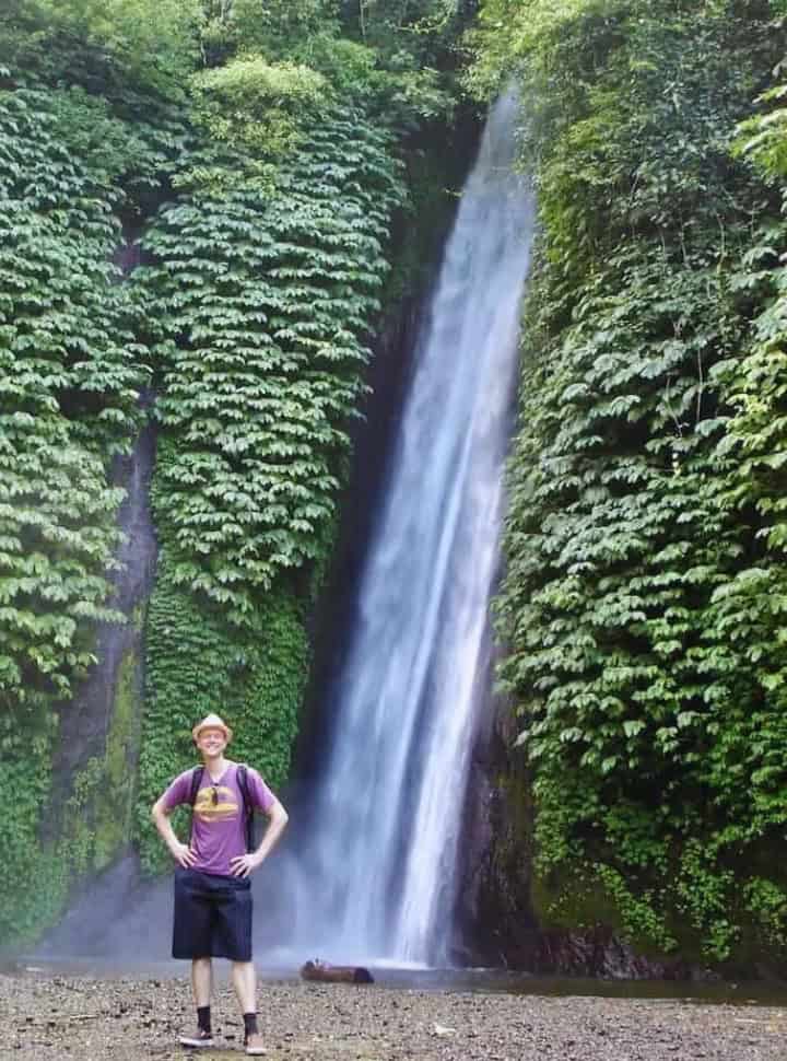 man posing in front of a waterfall rushing down into a pool with on both sides greenery, one of the waterfalls in munduk