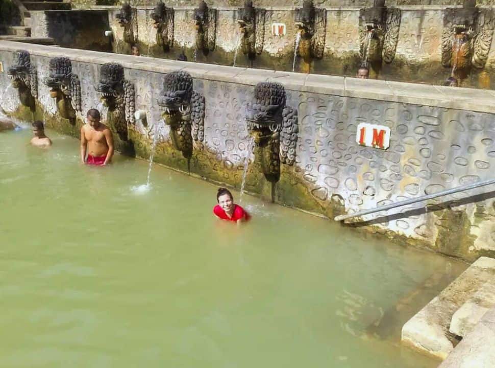 woman in a hot spring bath standing below a fountain of a balinese figure which is oozing water on her back 