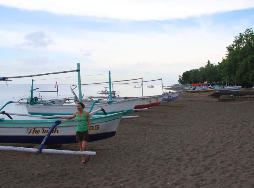woman standing on a dark colored sanded beach leaning against one of the many traditional wooden fishing boats at Lovina Beach Bali
