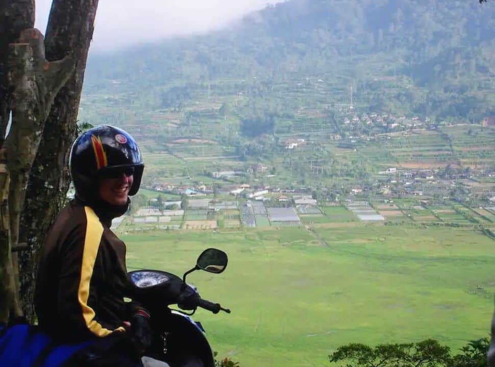 man on a motor scooter overlooking a green valley with houses and mountains nearby Bedugul Bali and Munduk