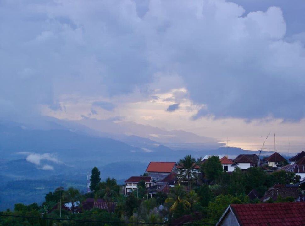 heave clouds hoovering over Munduk village and all the way into the mountain valley, a bit of sunlight is visible along the coast line in the far distance .