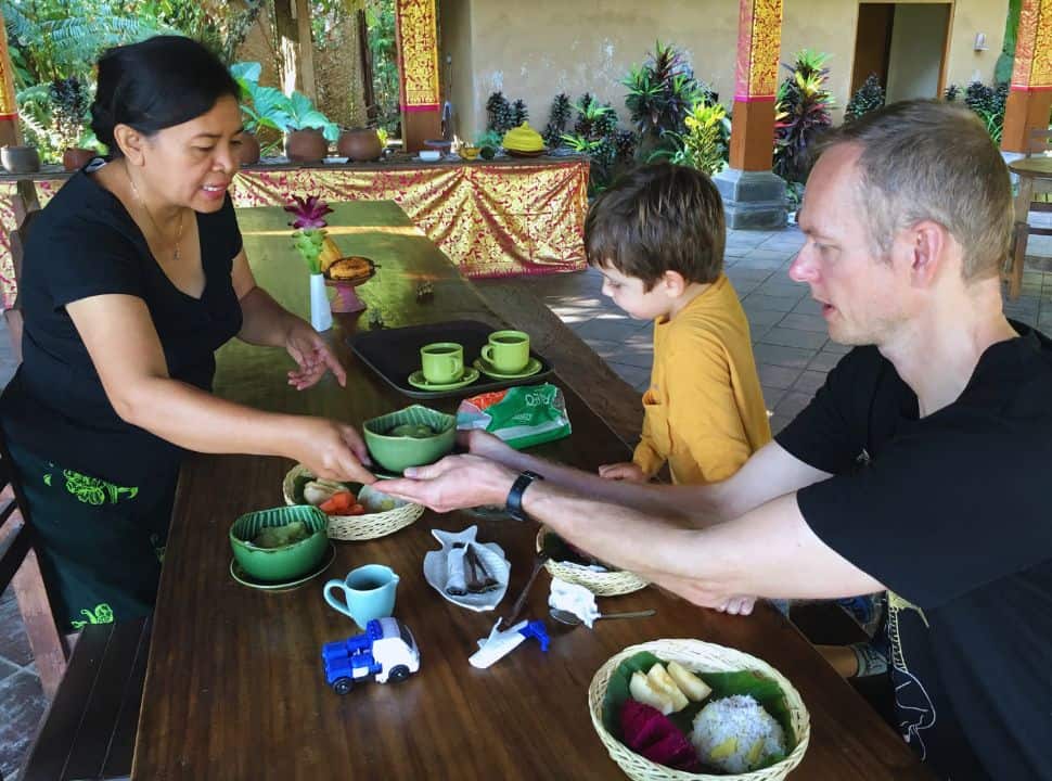 Balinese owner serving breakfast made from local ingredients to a father and son sitting at the table at their accommodation in Batukaru Bali