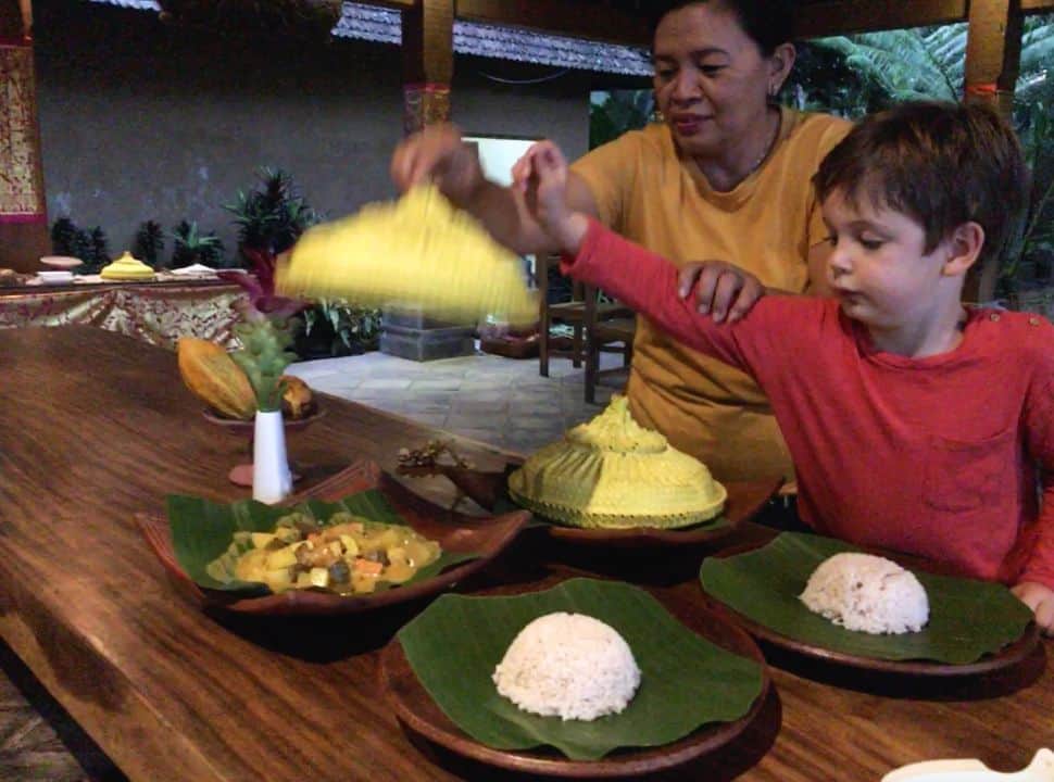 Balinese woman serving dinner, by lifting up a lid under which the food is placed. A little boy is checking out the food at Batukura accommodation in Bali