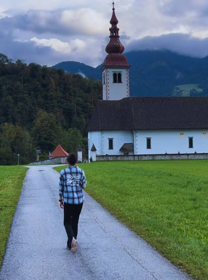 woman walking along a small road in Slovenia leading to a church with stunning mountain range in the back