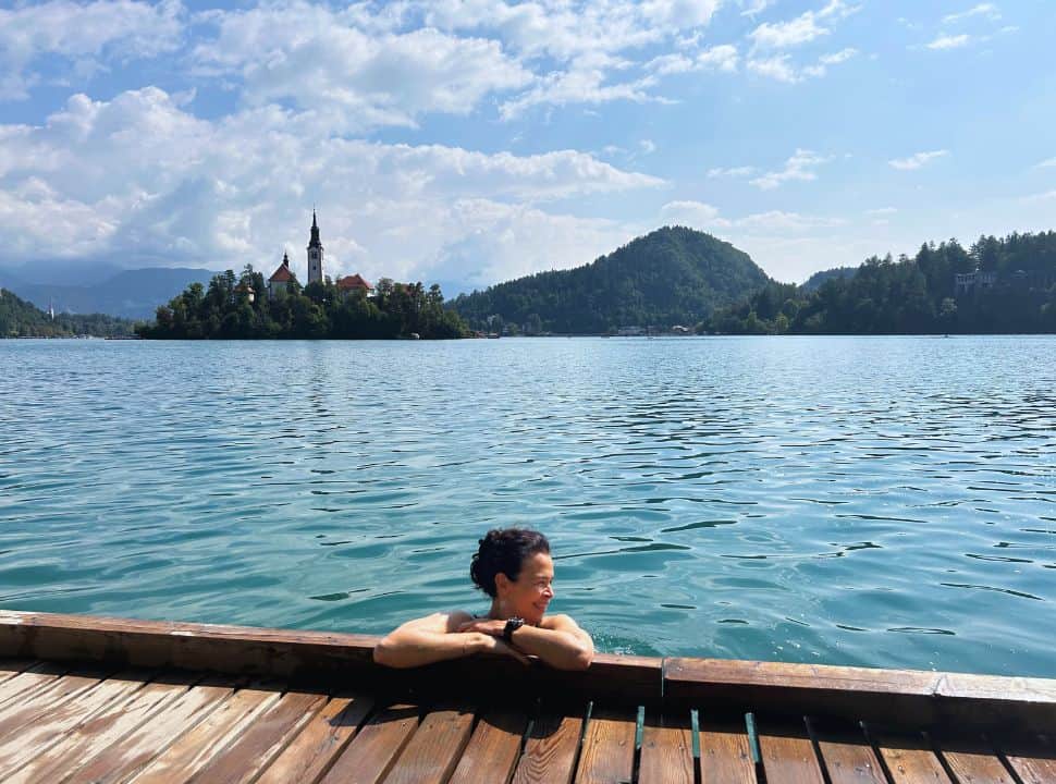 woman hanging from the water on the wooden walk way along the lake, enjoying the sun while in the back the famous Bled island with its church is clearly visible 