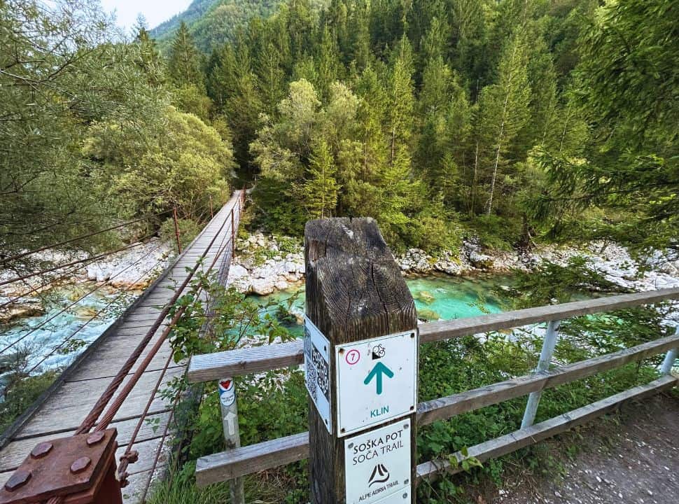 a sign pointing the trail across a suspension bridge that lies over a stunning emerald green river leading into the forest in Slovenia, Soca Valley