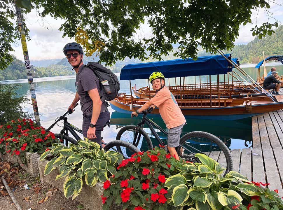 father and son both on a mountain bike posing on the docks at Lake Bled with in the background traditional boats