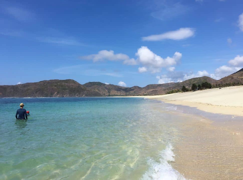 father and son swimming the the clear blue calm ocean water with view of a mountain range and stunning soft white long stretched beach in Lombok
