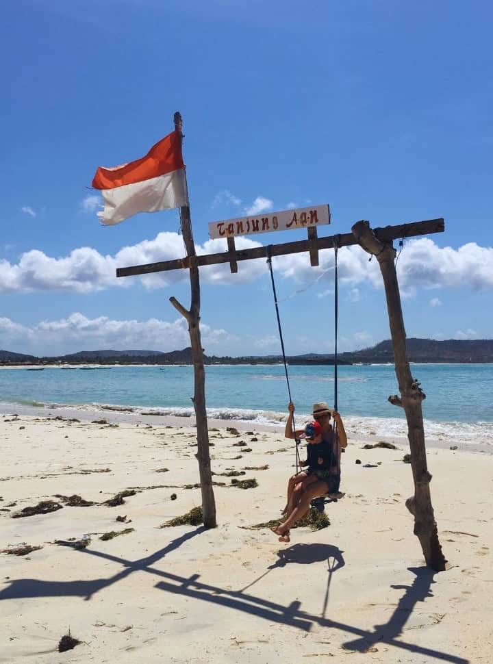 mother and son sitting on a wooden swing with the sign Tanjung Aan and the Indonesian Flag on a white sanded beach with stunning blue water in the back on Lombok Island. 