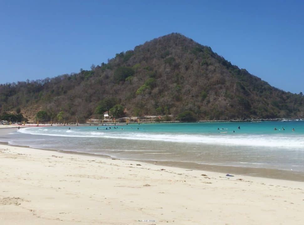 surfers enjoying the mild waves at the white sanded beach with turquoise blue water, a mountain hill dominating in the back in Lombok