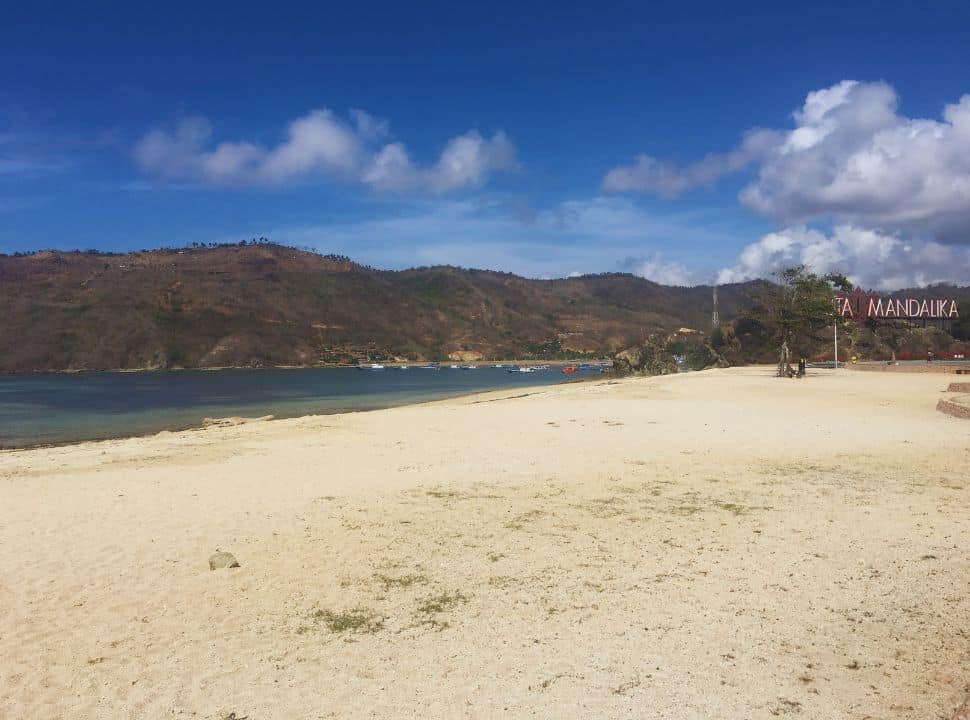 quiet beach with blue water, local fishing boats in the water and in the back dry hill slopes set against the blue sky at Kuta Mandalika Lombok