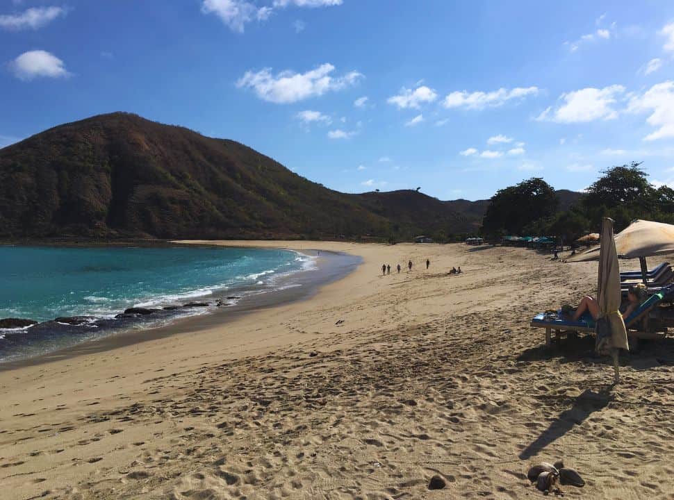 people walking a long stretched beach, sun beds are lined along the beach too. A hill is set in the distance at one of the beaches in Lombok 