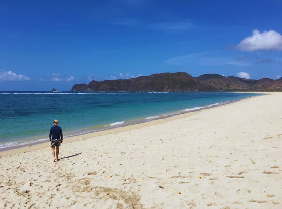 man walking on deserted long stretched beach in lombok with white soft sand, water is clear blue and in the distance there are bare hills