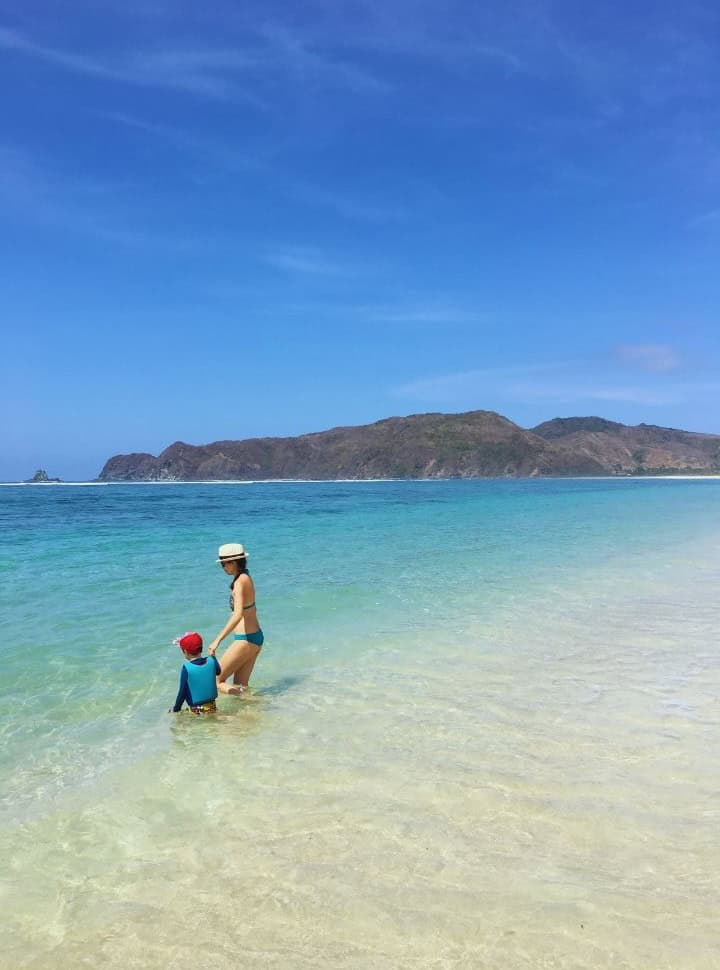 mother and son walking into the deep calm clear ocean water that has various colors blue at Tampah beach lombok, in the back are hills