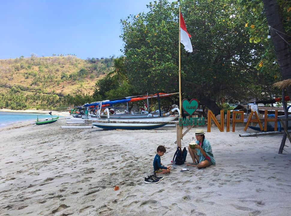 mother and son sitting on the beach drinking fresh coconet next to a I love pantai nipah sign, local fishing boats are lined just behind them.