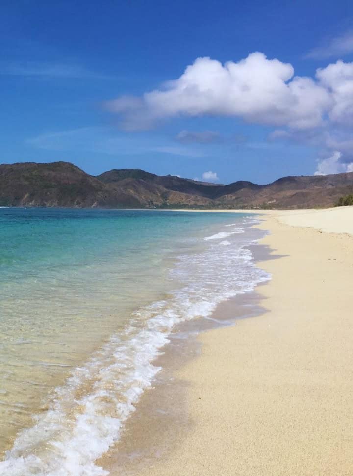 ocean shore line with water hitting the quiet soft sanded beach, blue water and hills in the back Lombok