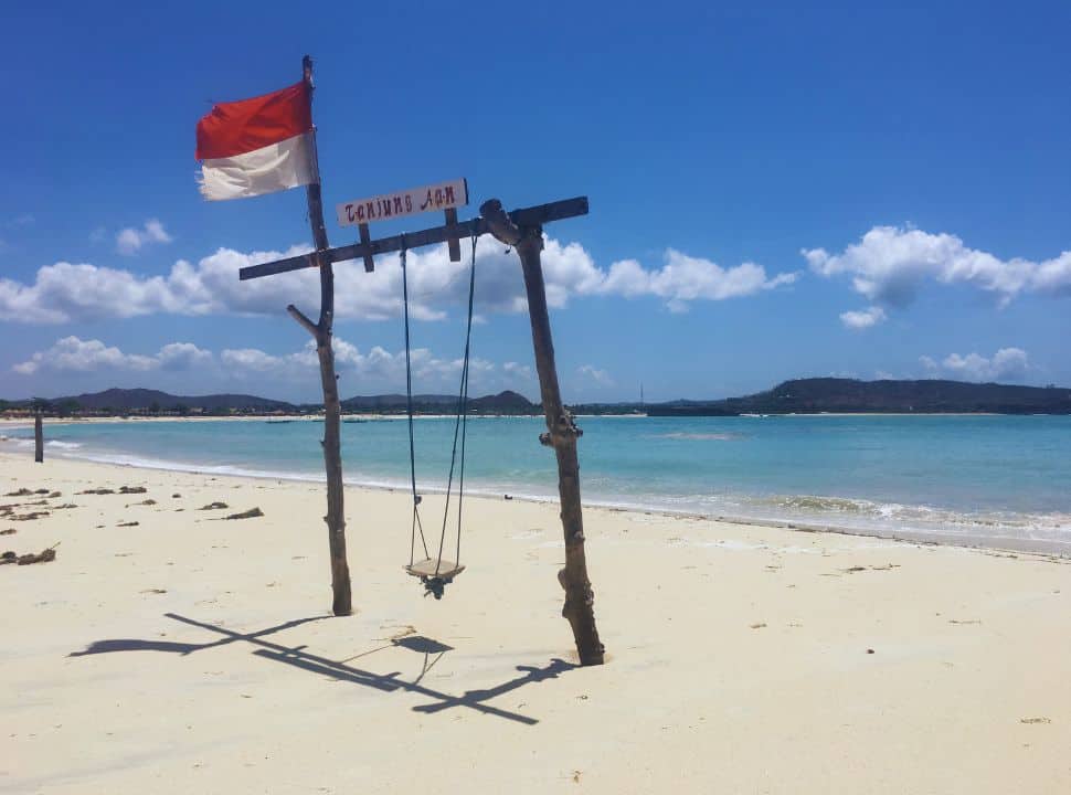 a wooden swing set on a soft white sanded beach with the sign Tanjung Aan and the Indonesian flag. In the back the ocean water is stunning blue
