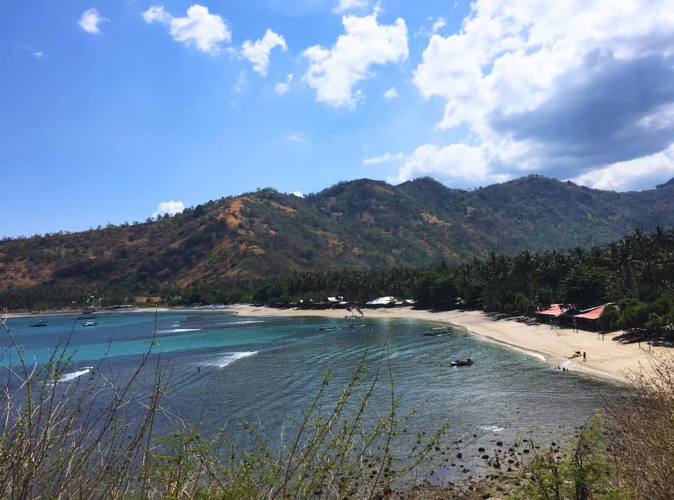 view of a strip of beach set between the hills, palm trees along the white sand. There are a couple of buildings visible and people swimming in the clear blue water near by Senggigi Lombok