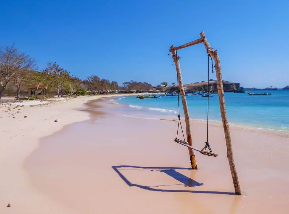 wooden swing set in sand with a slightly pink glow near the blue ocean water, trees line up along the beach while small boats are nearby the shore at pink beach Lombok