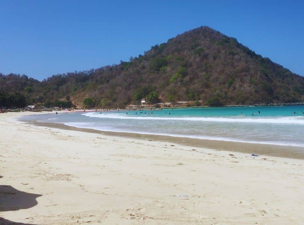 surfers waiting for a wave at a long stretched beach, water is turquise and the beach is white, a big hill closes the bay at Selong Belanak beach Lombok
