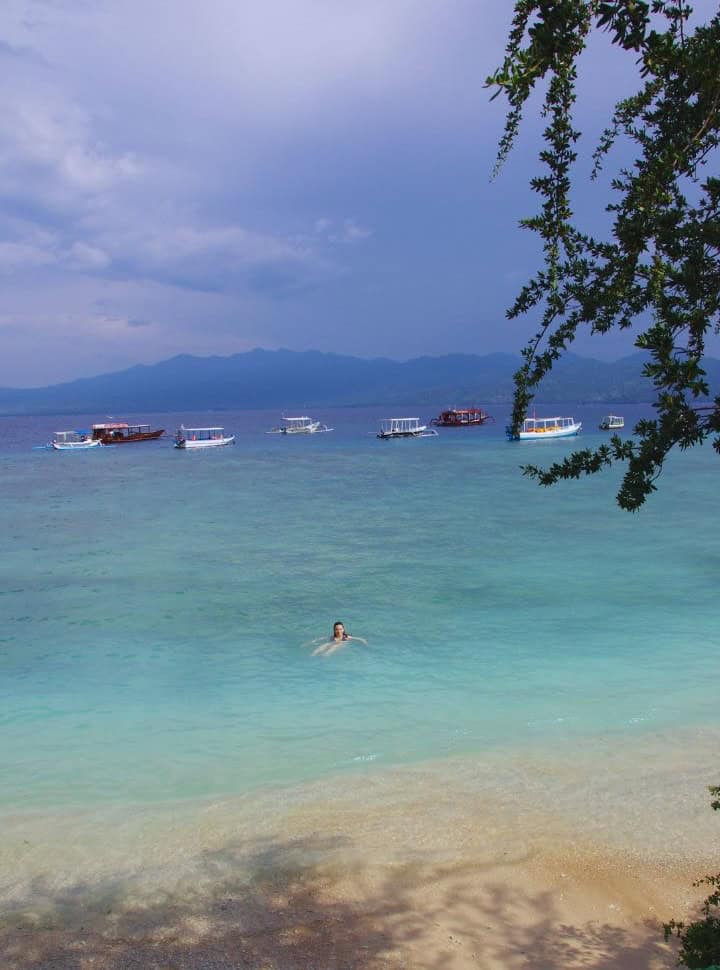 woman floating in the milky blue water just off the beach with in the back several traditional boats at Gili Trawangan Lombok