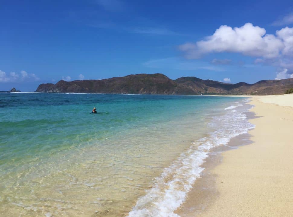long stretched tropical beach with white sand, two people are swimming in the clear water in shades of blue and turquoise, there is nobody else there, this beach in Lombok is deserted