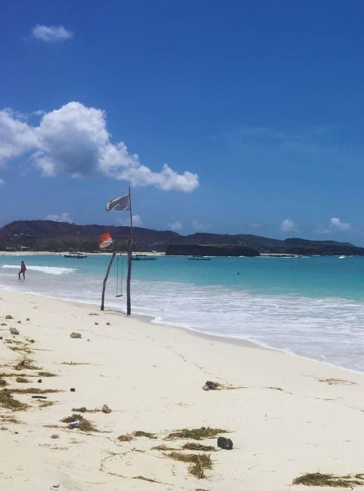 stunning soft white sand at Tanjung Aan beach Lombok where a swing is placed near the ocean front where the water is stunning blue, fishing boats are visible in the distance. 
