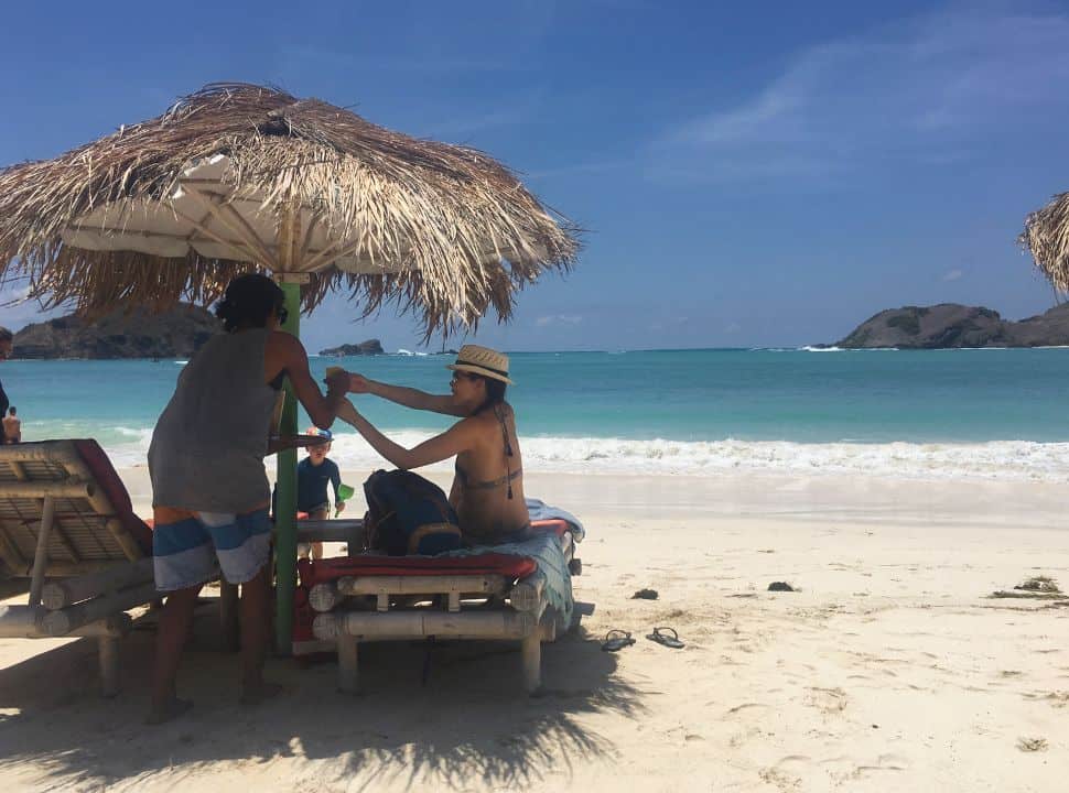 man handing a drink to a woman who is sitting under a sun umbrella and sitting on a bamboo sun lounger on a white sanded beach and clear blue ocean water at Tanjung Aa.