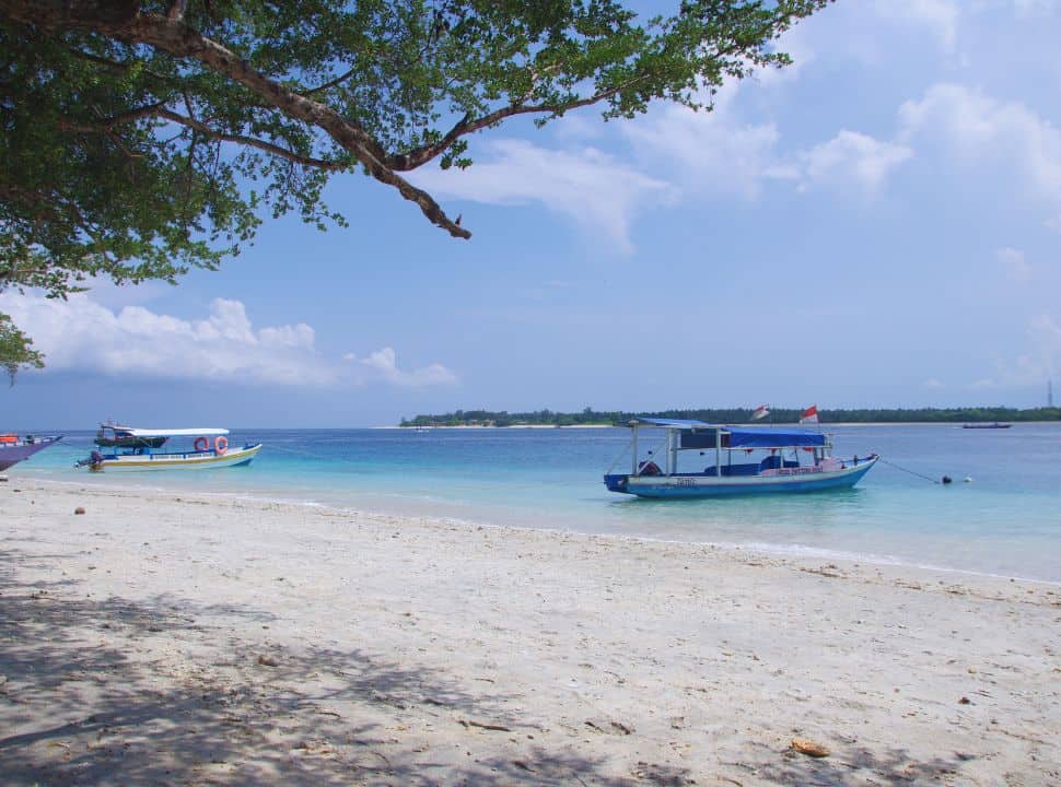 fishing boats just at the shore of a white sanded beach, in the distance a island is visible just across the stunning blue water at the Gili Island Lombok