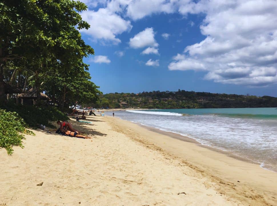 soft sanded beach with trees on one side providing shade and the ocean water on the other, people enjoying Jimbaran beach in South Bali