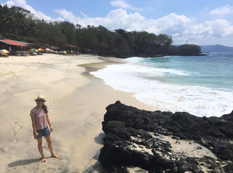 woman standing on a small deserted white sanded beach, volcanic rocks on the side with small beach shacks and sun umbrellas in east Bali