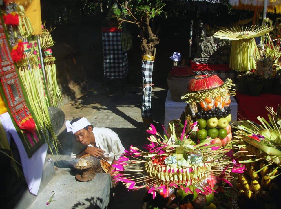 Balinese man in white blowing in a small offering creating some smoke, surrounded by colorful offerings in one of the many temples in Bali