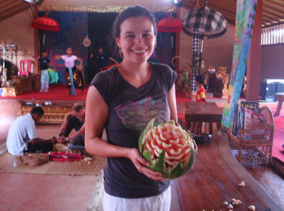 woman holding a watermelon carved into a flower, in the back young girls are practicing Balinese dancers and two men are wood carving in Ubud Bali