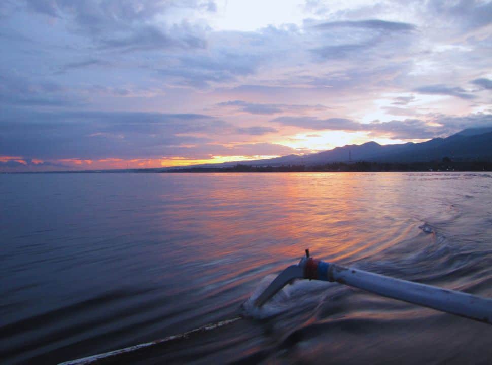 stunning colors in the sky and in the reflection of the ocean water while a traditional boat heads out from Lovina Bali. The mountains along the coast is still visible 